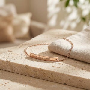 Op de afbeelding: Een roségouden armband met het woord "Unstoppable" gegraveerd op een gebogen staaf. De armband ligt op een getextureerd, lichtgekleurd oppervlak, met een zachte, gebroken witte doek op de achtergrond.