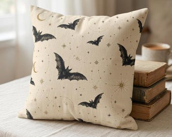 May include: A square throw pillow with a cream-colored fabric base featuring a repeating pattern of black bats, gold crescent moons, and various gold star shapes connected by thin, dotted lines across the entire surface of the pillow.