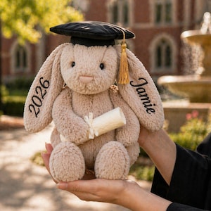 Puede incluir: Un conejito de peluche con birrete negro y borla dorada. El conejito sostiene un diploma enrollado y tiene bordados "2026" y "Jamie" en sus orejas. El conejito es de color marr&oacute;n claro.