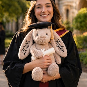 Puede incluir: Una graduada sonriente con birrete y toga negros sostiene un conejo de peluche con un gorro de graduaci&oacute;n en miniatura. El conejo beige tiene bordado "2026" y "Taylor". La graduada lleva una estola granate y dorada.