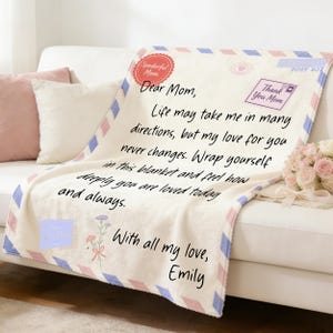 Puede incluir: Manta color crema con diseño de carta, con texto que dice "Querida mamá, la vida puede llevarme en muchas direcciones, pero mi amor por ti nunca cambia". Acentuada con patrones de diamantes rosas y azules.