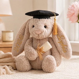 Puede incluir: Un conejito de peluche con birrete negro y borla dorada. El conejito es de color marr&oacute;n claro con texto bordado en las orejas que dice "2026" y "Sophia". Sostiene un diploma enrollado atado con una cinta dorada.