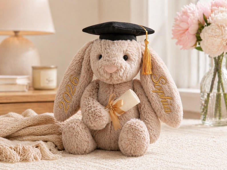 Puede incluir: Un conejito de peluche con birrete negro y borla dorada. El conejito es de color marr&oacute;n claro con texto bordado en las orejas que dice "2026" y "Sophia". Sostiene un diploma enrollado atado con una cinta dorada.