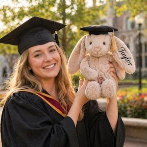 Puede incluir: Una graduada sonriente con birrete y toga negros sostiene un conejito de peluche con un gorro de graduaci&oacute;n en miniatura. El conejito es beige y tiene el nombre "Taylor" bordado en la oreja. El fondo muestra un campus universitario.