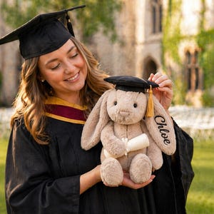 Puede incluir: Un conejito de peluche con un gorro de graduaci&oacute;n en miniatura y un diploma. La oreja del conejito est&aacute; bordada con el nombre "Chloe". El conejito es de color marr&oacute;n claro con gorro negro y borla dorada. La persona lleva una toga de graduaci&oacute;n.