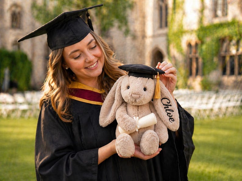 Puede incluir: Un conejito de peluche con un gorro de graduaci&oacute;n en miniatura y un diploma. La oreja del conejito est&aacute; bordada con el nombre "Chloe". El conejito es de color marr&oacute;n claro con gorro negro y borla dorada. La persona lleva una toga de graduaci&oacute;n.