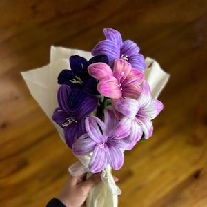May include: A bouquet of handmade fabric flowers in shades of purple, pink, and white, wrapped in cream-colored paper. The flowers are held together with a cream ribbon, creating a vibrant and colorful arrangement.