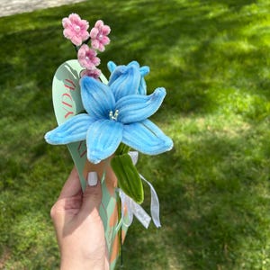 Op de afbeelding: Een handgemaakt bloemenboeket met een grote, helderblauwe bloem en kleinere roze bloemen. De bloemen zijn bevestigd aan een groene stengel en worden vastgehouden met een decoratieve kaart. Wit lint voegt een delicate touch toe.