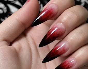May include: Stiletto-shaped press-on nails featuring a gradient design that transitions from a clear, translucent base at the cuticle to a deep, smoky red and black ombre effect at the sharp, pointed tips, creating a dramatic and bold aesthetic.