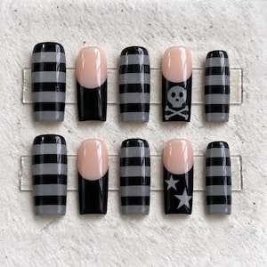May include: A set of ten press-on nails featuring a square shape. Some nails have horizontal black and gray stripes, while others feature a nude base with black tips. One nail displays a gray skull and crossbones, and another shows two gray stars.