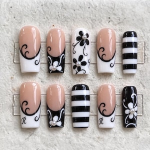 Puede incluir: Un juego de diez uñas postizas con una mezcla de diseños que incluyen bases color nude con patrones de remolinos negros, motivos florales blancos con pequeñas perlas, rayas horizontales blancas y negras, y la letra R en cursiva negra.