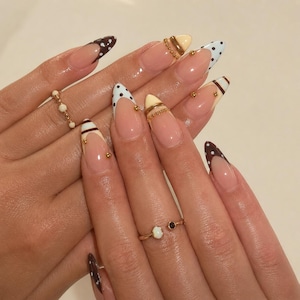 Puede incluir: Uñas en forma de almendra con una manicura francesa de diseños variados, incluyendo lunares marrones y blancos, puntas de color crema sólido y rayas marrones. El arte de uñas está acentuado con pequeñas cuentas y tachuelas doradas.
