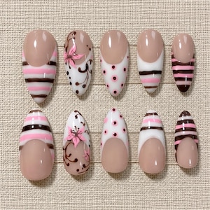 May include: A set of ten almond-shaped press-on nails featuring a nude base with various designs including pink and brown horizontal stripes, white backgrounds with pink polka dots, and white backgrounds with hand-painted pink floral motifs.