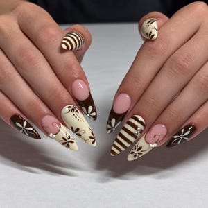 May include: Almond-shaped nails featuring a mix of dark brown and cream polish. Designs include brown and cream horizontal stripes, intricate hand-painted white floral patterns with brown centres, and classic French tips in dark brown.