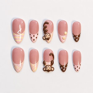 May include: A set of ten almond-shaped press-on nails featuring a nude base with various designs including brown and white polka dots, yellow and white plaid patterns, yellow stripes, and two nails with raised 3D brown monkey character accents.