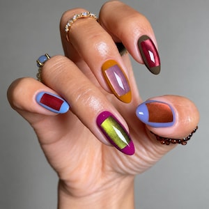 Puede incluir: Uñas de forma ovalada con una manicura multicolor. Cada uña tiene un color base sólido con un centro rectangular contrastante con acabado metálico o efecto ojo de gato. Los colores incluyen amarillo mostaza, marrón oscuro, magenta y azul claro.