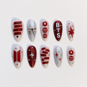 Pode incluir: Um conjunto de dez unhas postiças em formato amendoado com uma paleta de cores vermelho, branco e prata. Os designs incluem o texto ARMY YOU GOT MY BACK, um laço prateado, as letras BTS, motivos de estrelas, o logótipo BTS e os nomes.