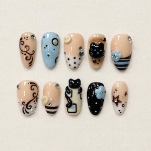 May include: A set of ten press-on nails featuring a variety of designs including nude, light blue, and black base colors. The nails are decorated with black swirls, polka dots, horizontal stripes, stars, and three-dimensional black cat figures.