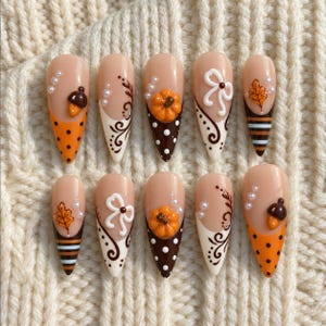 Op de afbeelding: Een set van tien amandelvormige kunstnagels met een nude basis en herfstthema-ontwerpen, waaronder oranje en bruine stippen, witte krullen, 3D oranje pompoenen, witte strikken, bruine en witte strepen, oranje bladeren en kleine witte parels.