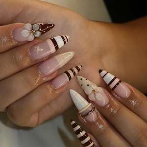 Op de afbeelding: Amandelvormige nagels met een mix van ontwerpen, waaronder bruine en witte strepen, crèmekleurige stippen, een ruitpatroon en driedimensionale witte bloemaccenten met kleine gouden hartjes. Sommige nagels hebben een doorzichtige basis met een ster.