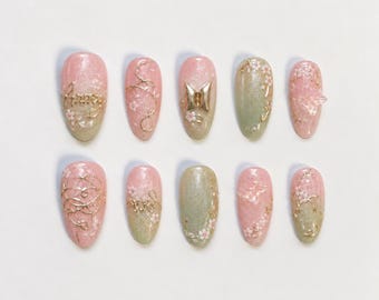 Peut inclure: Dix faux ongles avec un dégradé de rose tendre et de vert sauge à la finition scintillante. Les ongles sont décorés de motifs de fleurs de cerisier blanches, de fils dorés et d'un logo métallique doré. Un ongle porte le mot ARMY en doré.