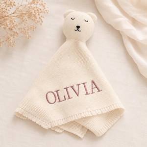 May include: A cream-colored knitted security blanket featuring a bear head with embroidered closed eyes and a nose. The fabric has a textured knit pattern and includes the name OLIVIA embroidered in a dusty rose thread across the center.