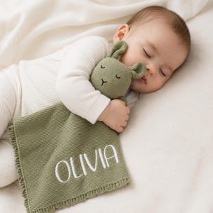 May include: A sage green knitted security blanket featuring a plush rabbit head with embroidered closed eyes and a small nose. The square blanket has a textured knit border and the name OLIVIA is embroidered in white capital letters.