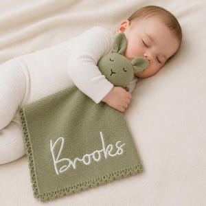 May include: A sage green knitted security blanket featuring a plush rabbit head with embroidered closed eyes and a nose. The blanket has a decorative scalloped edge and the name Brooks is embroidered in white cursive script on the front surface.