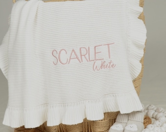 Puede incluir: Una manta de punto blanca con borde de volantes, que presenta el texto bordado SCARLET White en hilo rosa claro. La manta está colocada sobre una cesta de mimbre tejida, acompañada por un par de pequeños patucos de punto con diseños de nubes.