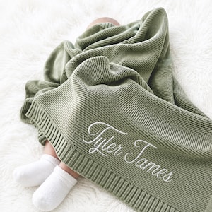 May include: A sage green knitted baby blanket featuring a ribbed border and the name Tyler James embroidered in white cursive script. The blanket has a textured garter stitch pattern and is draped over a pair of small feet wearing white socks.