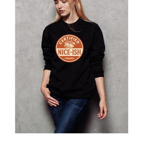 May include: Black sweatshirt featuring the text "CERTIFIED NICE-ISH" in a circular design. The design includes bells and decorative elements in orange and white. The model is wearing blue jeans.