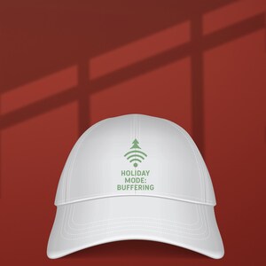 May include: A white baseball cap with a curved brim. The cap features a green graphic of a Christmas tree and Wi-Fi symbol, with the text "HOLIDAY MODE: BUFFERING" in green. The background is a solid red.
