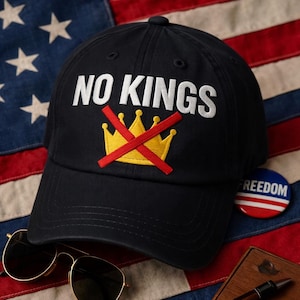 May include: A black baseball cap with white embroidered text reading "NO KINGS" above a yellow crown crossed out with a red line. A "FREEDOM" badge and sunglasses are also visible.
