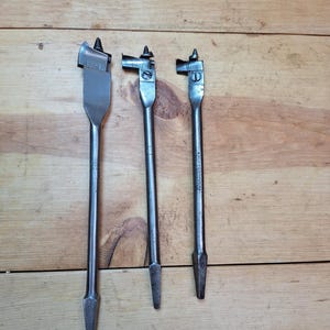 May include: Three silver-colored spade drill bits of varying sizes, arranged vertically against a wooden background. The bits have a flat, paddle-shaped cutting edge and a pointed tip. The wood grain is visible.