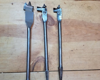 May include: Three silver-colored spade drill bits of varying sizes, arranged vertically against a wooden background. The bits have a flat, paddle-shaped cutting edge and a pointed tip. The wood grain is visible.