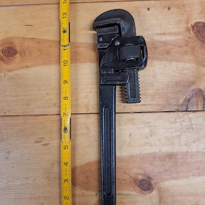 May include: A black pipe wrench with adjustable jaws, next to a yellow and black measuring tape. The wrench is approximately 12 inches long. The wrench is on a wooden surface.