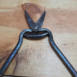 May include: Close-up of antique metal shears with pointed blades and curved handles. The shears are dark gray with signs of wear, suggesting age and use. The blades are open, revealing the cutting edges. The shears are resting on a wooden surface.