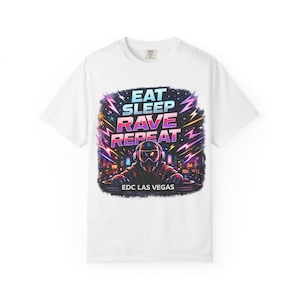 Puede incluir: Camiseta blanca con un diseño gráfico colorido. El diseño presenta el texto "EAT SLEEP RAVE REPEAT" sobre una ilustración de un DJ con auriculares, con el texto "EDC LAS VEGAS" debajo. El diseño incluye rayos y un paisaje urbano.