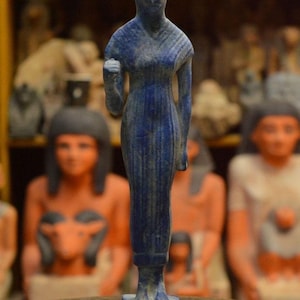 May include: A blue statue of the Egyptian goddess Bastet, depicted with a cat's head and a slender, standing form. The statue is on a rectangular base with carved details. Other statues are visible in the background.