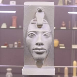 Peut inclure: Sculpture rectangulaire blanche représentant le visage d'un pharaon, orné d'une coiffe avec un cobra. La sculpture est exposée dans une vitrine avec d'autres objets.