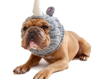 May include: A tan bulldog wearing a gray knit unicorn hat with a white horn and pink inner ears. The dog is lying down on a white background, looking directly at the camera.