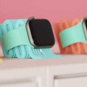 May include: Three smartwatches with black screens and colourful bands are displayed on a white surface. The bands are in shades of yellow, teal, and coral, each resting on a matching stand. The background is a soft pink.