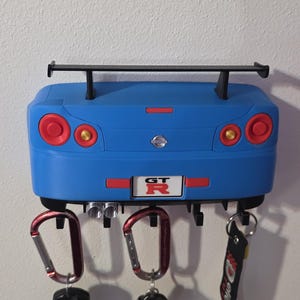 Puede incluir: Un llavero azul con forma de coche, con alerón y el texto "GT R". Tiene luces traseras rojas y sujeta llaves con mosquetones.