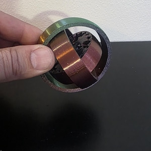 May include: A 3D-printed puzzle with a circular frame in green and purple hues, surrounding intersecting copper-colored bands. The bands enclose a dark, textured core. The design is held by a hand.