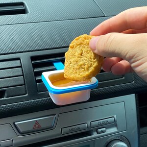 May include: A golden-brown chicken nugget being dipped into a small, white and blue sauce container. The container is attached to a car's air vent. The car's dashboard is visible in the background.