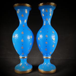 Puede incluir: Dos jarrones de cristal azul a juego con diseños florales dorados y bordes dorados. Los jarrones tienen un cuerpo bulboso y una parte superior e inferior ensanchadas. El diseño floral dorado está distribuido por la superficie de los jarrones.