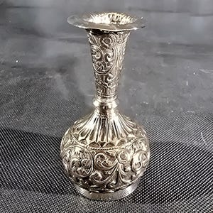 May include: A silver-colored vase with intricate floral and scrollwork patterns. The vase has a flared top, a slender neck, and a rounded base. The detailed design covers the entire surface of the vase, creating a textured appearance.