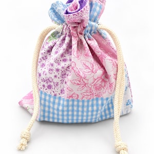 May include: A small fabric drawstring pouch featuring a patchwork design with various floral patterns in shades of pink and purple, combined with a light blue and white gingham check fabric at the base. The bag is closed with two thick cream cords.