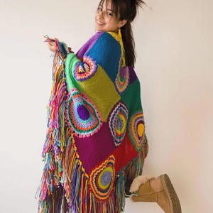May include: A vibrant, multi-coloured crochet poncho featuring a patchwork design of solid squares and circular motifs in shades of blue, green, purple, red, and yellow. The garment is finished with long, thick, multi-coloured yarn fringe trim.