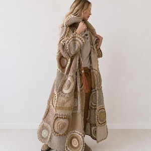 Könnte beinhalten: Ein langer, vorne offener Häkelmantel mit einem Patchwork-Design aus kreisförmigen und quadratischen Motiven in verschiedenen Beige-, Braun- und Ockertönen. Das Kleidungsstück verfügt über eine Kapuze, lange Ärmel und eine dekorative Quaste.